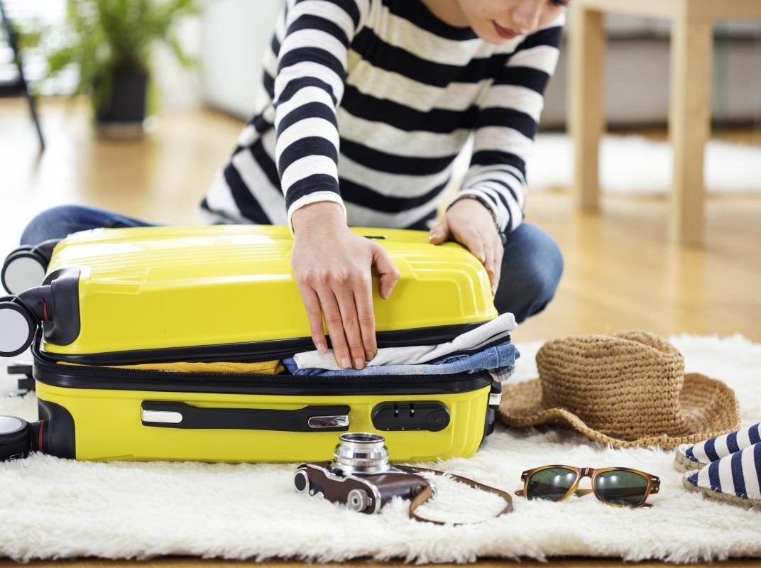 Person sitzt auf dem Boden, versucht gelben Koffer zu schließen. Drumherum liegen noch Reiseutensilien.
