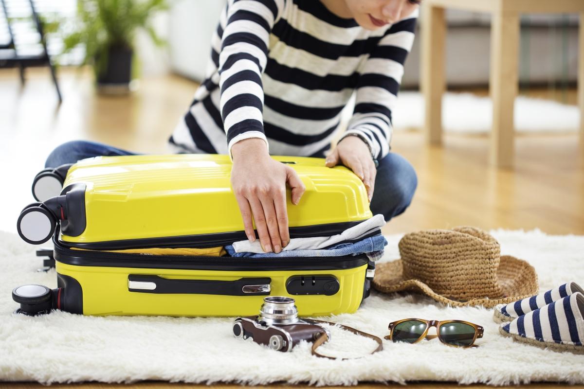 Person sitzt auf dem Boden, versucht gelben Koffer zu schließen. Drumherum liegen noch Reiseutensilien.